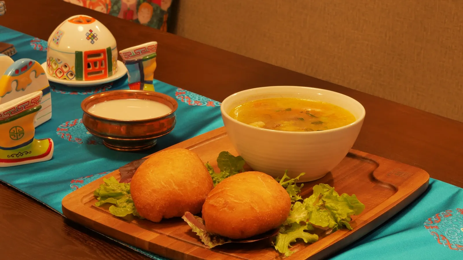 Piroshki and soup on a timber board beside a ger-shaped teapot and milk tea